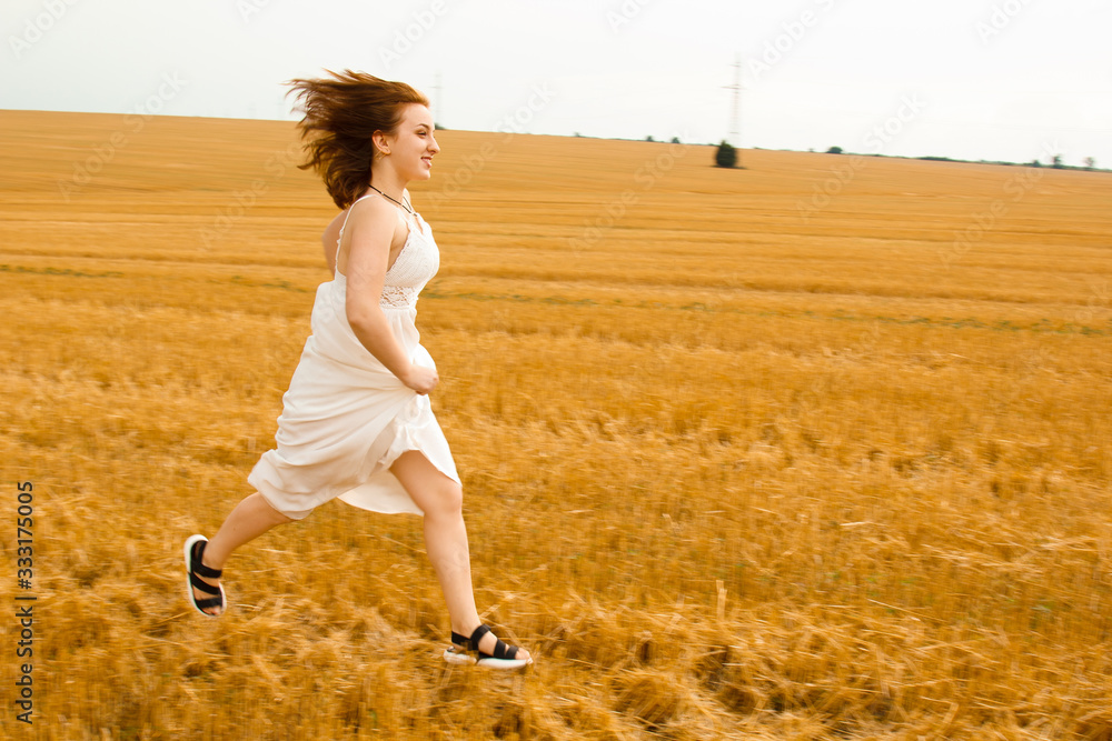 Runs across the field. Young beautiful redhead woman in the middle of a wheat field having fun. Summer landscape, good weather. Windy day with the sun and clouds. White cotton dress, eco style. Runs across the field. Young beautiful redhead woman in the middle of a wheat field having fun. Summer landscape, good weather. Windy day with the sun and clouds. White cotton dress, eco style.