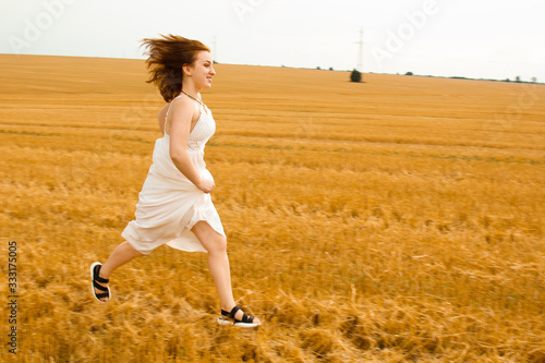 Runs across the field. Young beautiful redhead woman in the middle of a wheat field having fun. Summer landscape, good weather. Windy day with the sun and clouds. White cotton dress, eco style.