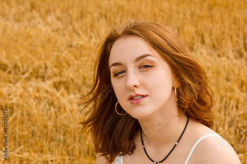 Portrait of a young beautiful redhead girl in the middle of a wheat field. He looks into the frame, brown eyes. Windy plays with hair. White cotton dress, eco style.