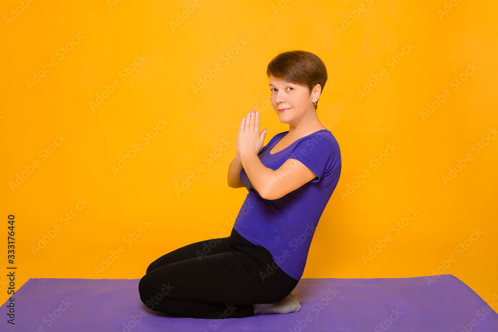 Fototapeta premium Aged woman doing yoga on a purple rug. Studio photo on a yellow background.