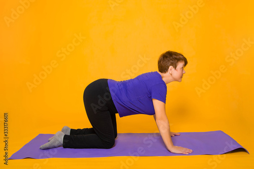 Aged woman doing yoga on a purple rug. Studio photo on a yellow background.