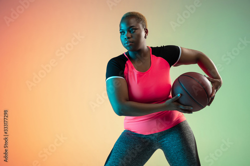 Young african-american plus size female model's training on gradient background in neon light. Doing workout exercises with the ball. Concept of sport, healthy lifestyle, body positive, equality. photo