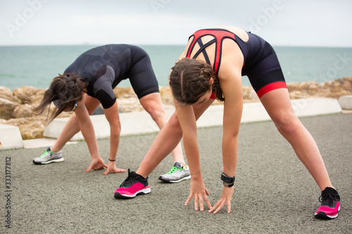 Sporty women stretching near sea. Full length view of athletic women in sportswear stretching and working out on sea coast. Triathlon concept
