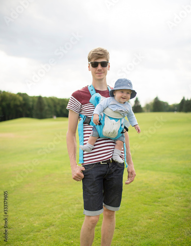 Happy young cool father with baby in sling in front having a walk outside in park or green meadow. Parenthood and daddy time. Posing on camera and smile.