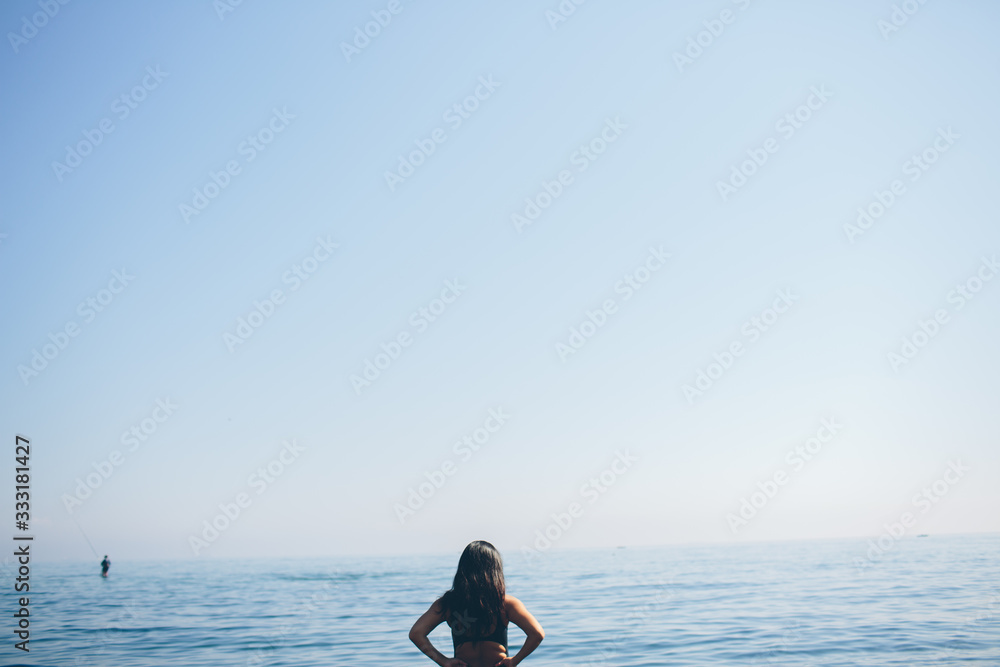 Young woman standing and looking at ocean and in sky. Female figure alone on beach. Blue sky and ocean water. Alone.