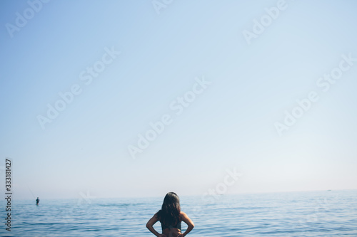 Young woman standing and looking at ocean and in sky. Female figure alone on beach. Blue sky and ocean water. Alone.