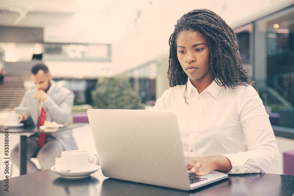 Serious female professional working on laptop while drinking coffee in office lobby. Businessman in formal suit using tablet in background. Workspace in cafe concept Serious female professional working on laptop while drinking coffee in office lobby. Businessman in formal suit using tablet in background. Workspace in cafe concept