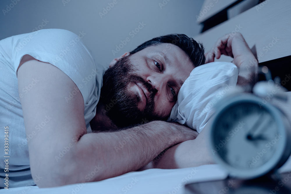 A lonely bearded man in bed at night suffers from insomnia and he watches the time on the alarm clock. Can not fall asleep, a clock on the pedestal, a bedroom, pajamas, white bedding. - obrazy, fototapety, plakaty 