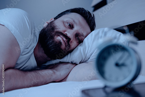 A lonely bearded man in bed at night suffers from insomnia and he watches the time on the alarm clock. Can not fall asleep, a clock on the pedestal, a bedroom, pajamas, white bedding.