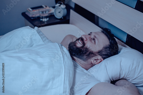 A lonely bearded dark-haired man with a very happy face lies on his back and looks at the ceiling at night on the bed. Dreams, a date succeeded, happy, white bedding.