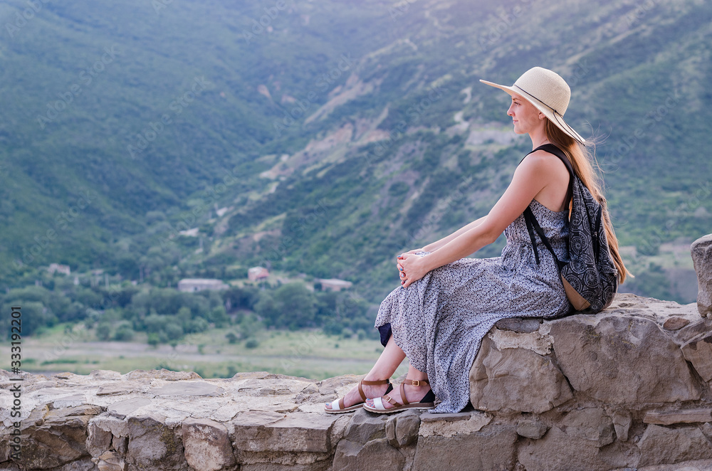 Naklejka premium Beautiful young woman in dress and hat sitting on background of mountain valley