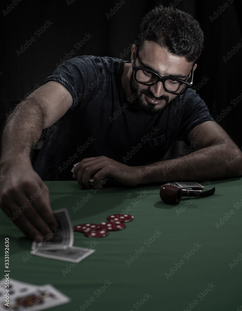 Portrait of young brunette Indian Kashmiri man in casual tee shirt playing cards on a casino poker table in black copy space studio background. lifestyle and fashion. Portrait of young brunette Indian Kashmiri man in casual tee shirt playing cards on a casino poker table in black copy space studio background. lifestyle and fashion.