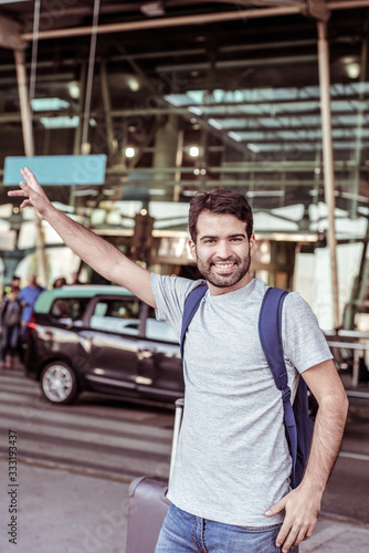 Front view of smiling traveler waving hand near airport. Cheerful young man trying to stop car. Travel concept