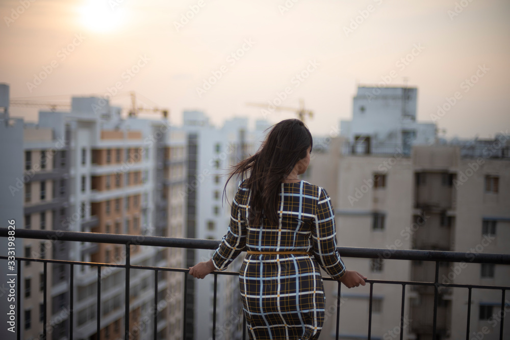 Back side of an young brunette Indian Bengali brunette plus size woman in western dress standing on rooftop in urban background while her hair is blowing in wind during sunset. lifestyle and fashion.