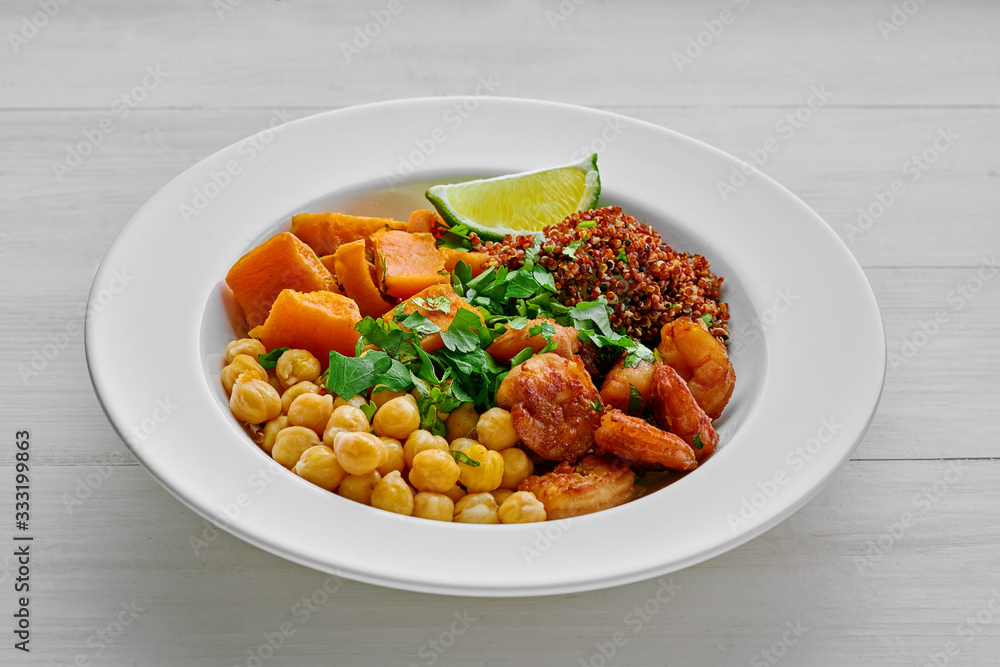 Delicious bowl with garlic grilled shrimps, sweet potato, quinoa, chickpeas and lime on a white wooden table.