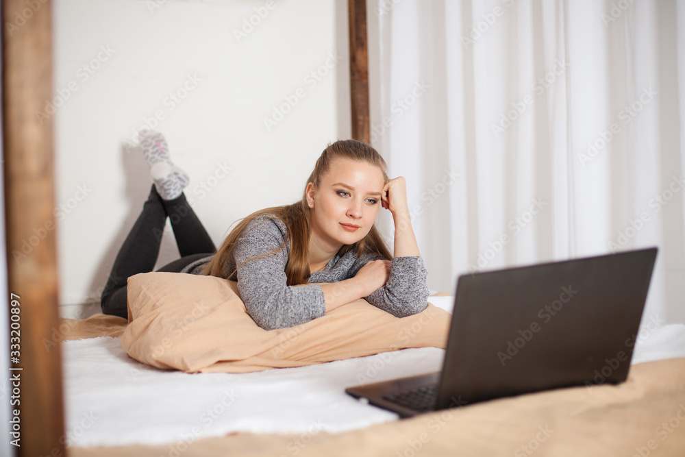  A beautiful girl with brown hair is lying on a bed with a laptop and watching a movie. A white canopy bed with brown linens.