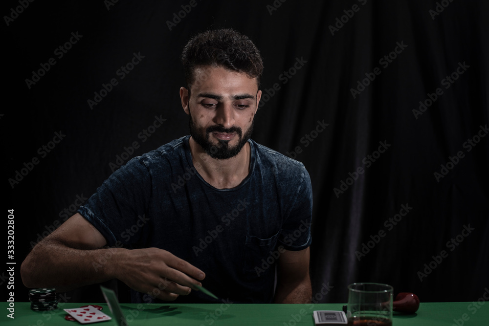 Portrait of young brunette Indian Kashmiri man in casual tee shirt playing cards on a casino poker table in black copy space studio background. lifestyle and fashion. Portrait of young brunette Indian Kashmiri man in casual tee shirt playing cards on a casino poker table in black copy space studio background. lifestyle and fashion.