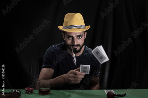 Portrait of young brunette Indian/European/Arabian/Kashmiri man in casual tee shirt and yellow hat playing cards on a casino poker table in black copy space studio background. lifestyle and fashion.