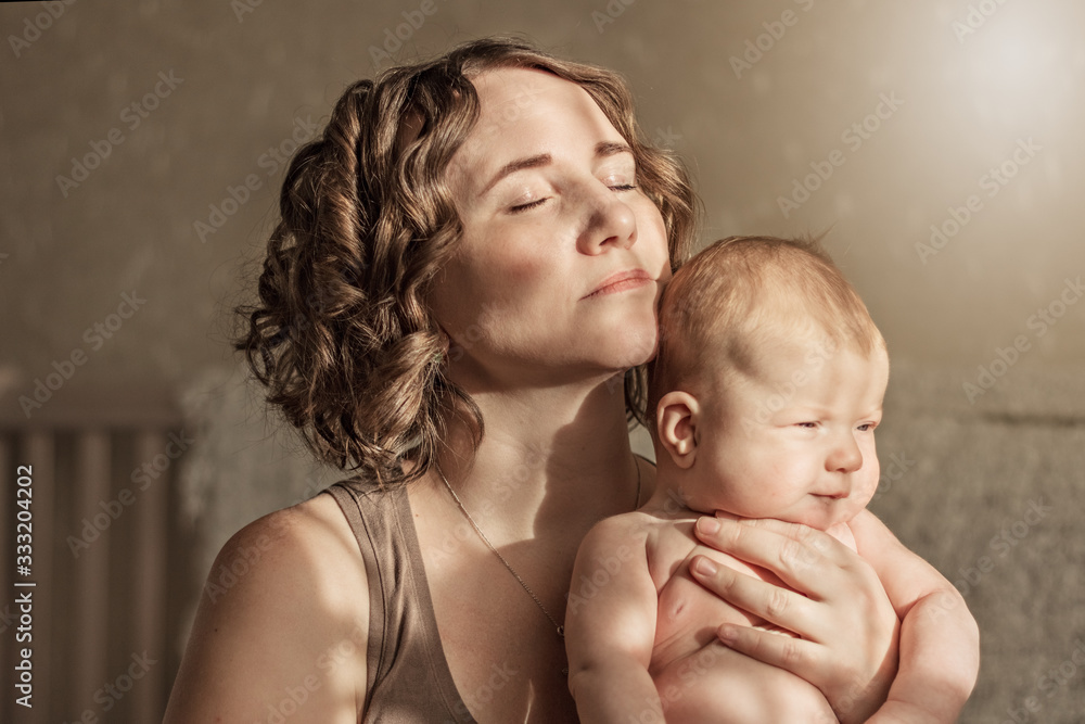 A mother with curly hair gently holds her little baby in her arms in the sun. Lifestyle at home with natural light.