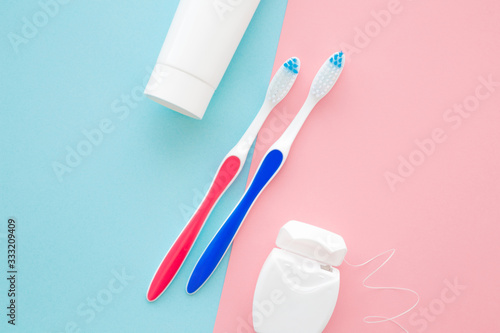 White tube of toothpaste, two toothbrushes and container of dental floss on pastel blue pink table background. Male and female teeth hygiene concept. Closeup. Top down view. Two sides.