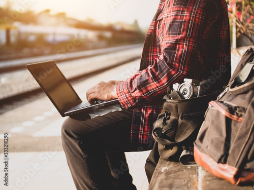 Summer day of Asian man using social media at train station. Travel concept. Hipster man hand using phone and laptop sitting with backpack and waiting for the train to go traveling.