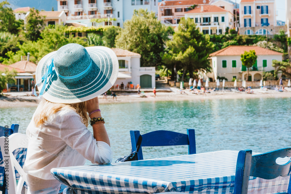 Obraz premium Rear view of tourist woman sitting in a cafe wear blue sunhat enjoying colorful tranquil houses of Assos village on sunny day. Kefalonia, Greece