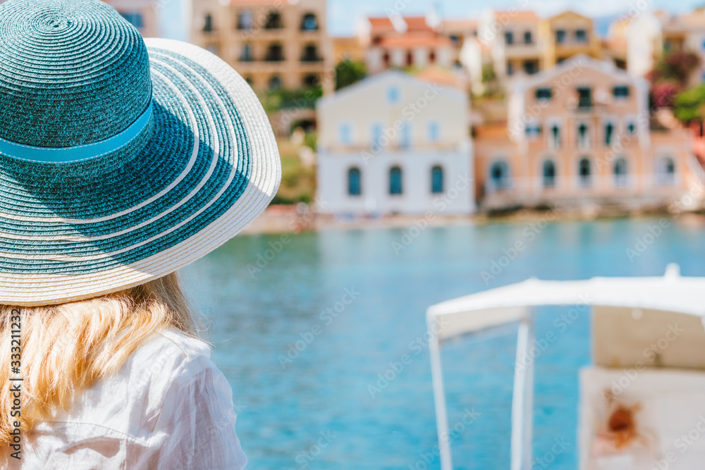 Rear close up view of tourist woman wear blue sunhat admire colorful tranquil houses of Assos village on sunny day. Visiting Kefalonia in summer. Travel vacation in Greece