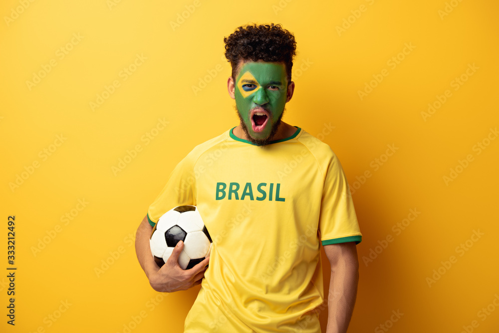 emotional african american football fan with face painted as brazilian flag yelling and holding ball on yellow emotional african american football fan with face painted as brazilian flag yelling and holding ball on yellow