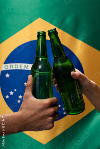 cropped view of couple of football fans clinking with bottles of beer with brazil flag behind