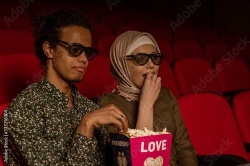 African american muslim man with his wife sitting in movie theater, watching 3D movie, eating popcorn, smiling.