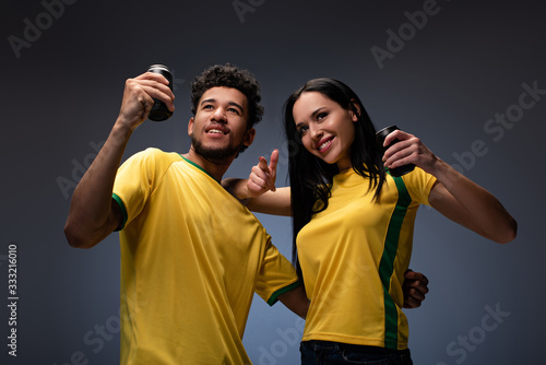 multiethnic couple of smiling football fans in yellow t-shirts holding cans with drink on grey photo