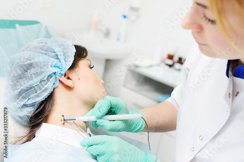 A young woman doctor dermatologist surgeon holds a radio wave knife in the background of a young female patient. Removal of moles, warts, rosacea, vascular asterisks, pigmentation.