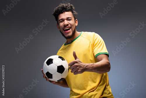 happy african american football fan in yellow t-shirt holding ball and showing thumb up on grey © LIGHTFIELD STUDIOS