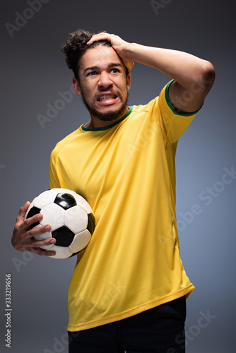 stressed african american football fan in yellow t-shirt holding ball on grey