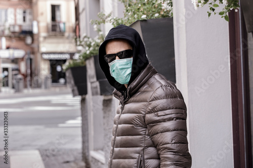 Ragazzo rasato con giubotto scuro ecappuccio in testa indossa una mascherina protettiva chirurgica mentre sta in giro per la città  photo