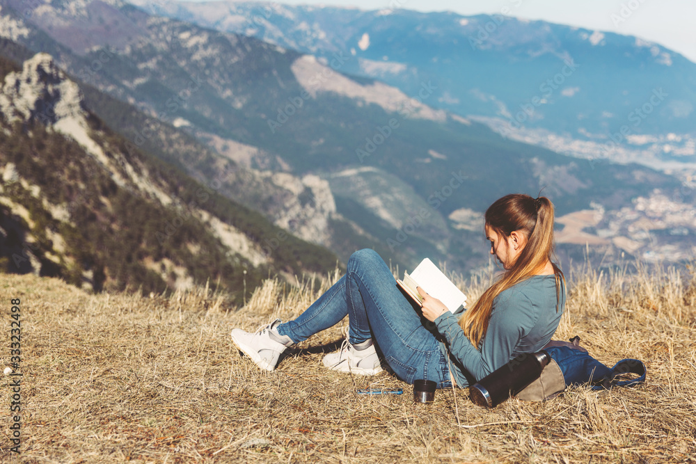 Young beautiful girl travels alone in the mountains in spring or autumn, sits on the edge of the mountain and reading book and enjoys nature, rocks and green forests, view of the landscape. a backpack