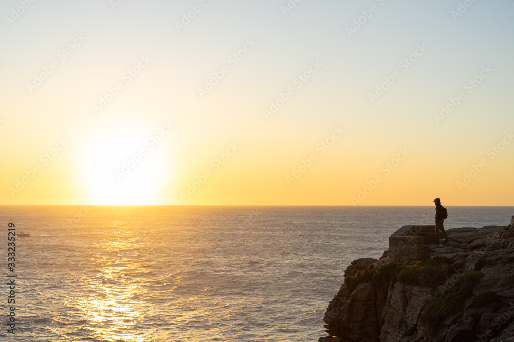 A person in the distance standing on the edge of a cliff looking at the ocean in the sunset. View from the side.