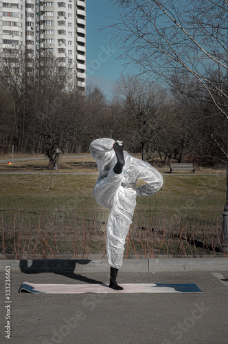 Life during epidemic. Woman in a protective suit, mask and gloves makes yoga exercises in empty city park. Summer season, selective focus. Lifestyle and sport during coronavirus (covid-19) pandemic.