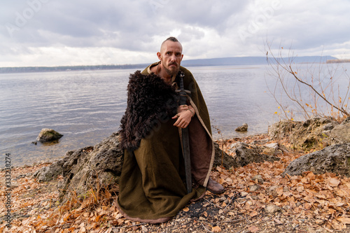 a viking dressed in a cape and skin armed with a sword and shield sits on a stone against the background of the river