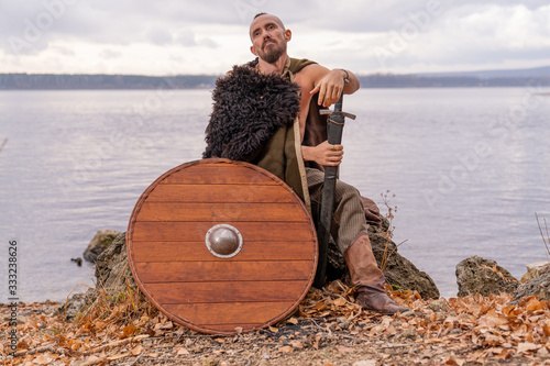 a viking dressed in a cape and skin armed with a sword and shield sits on a stone against the background of the river