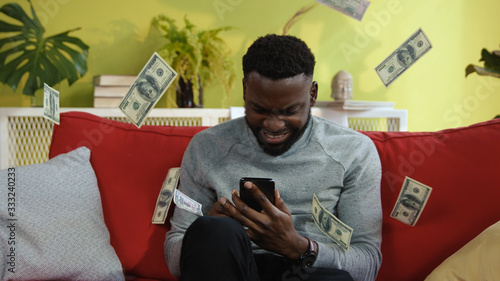 Happy african man winner reading exciting news on smartphone overjoyed with success online game win staying euphoric in cozy home room interior.