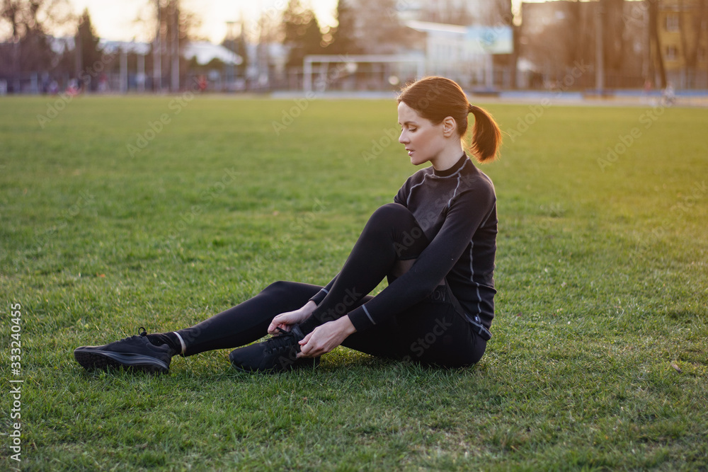 Sporty brunette fit young woman wearing black sport uniform tying shoelaces on sneakers sitting on a green lawn Sporty brunette fit young woman wearing black sport uniform tying shoelaces on sneakers sitting on a green lawn