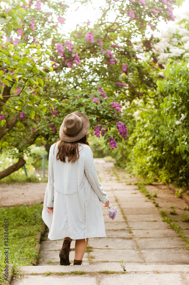 Stylish girl in a light coat and hat walks in the garden of a blossoming lilac Stylish girl in a light coat and hat walks in the garden of a blossoming lilac