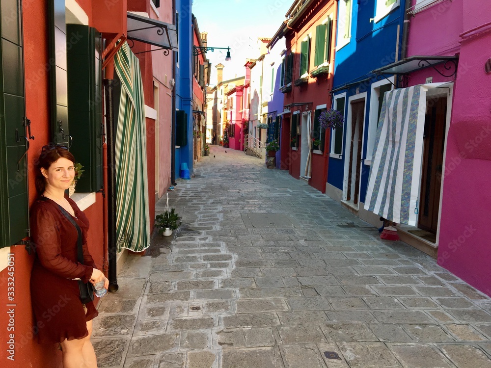 A beautiful young female tourist posing along a narrow brick road full of old colourful homes and flowers in Burano, Italy, an island outside of Venice.  The colours are shades of purple, red, blue.