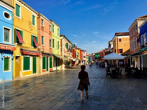 A view of the quiet town square of Burano, Italy during the early morning. A few people walk past the colourful shops with the leaning bell tower in the background.