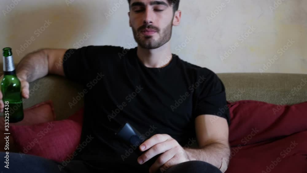 Young attractive man sitting on couch at home in living-room, drinking beer and eating snacks from a bowl, relaxing in a lazy afternoon Young attractive man sitting on couch at home in living-room, drinking beer and eating snacks from a bowl, relaxing in a lazy afternoon