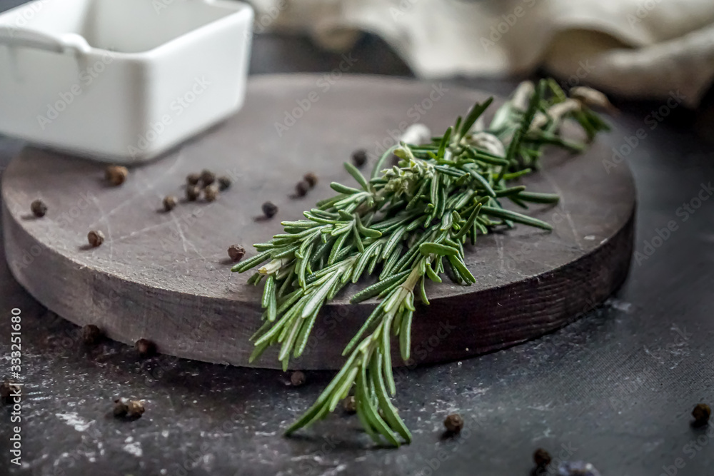 A bunch of rosemary and pepper in a white bowl on a wooden board on a background of dark concrete table and a beige towel. Herbs and spices.