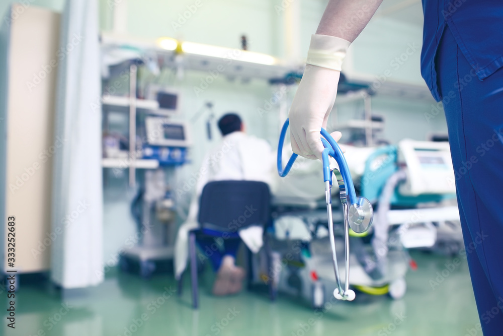 Fototapeta premium Figure of a doctor with stethoscope in hands on the background of a man siiting near the patient bed in hospital ward