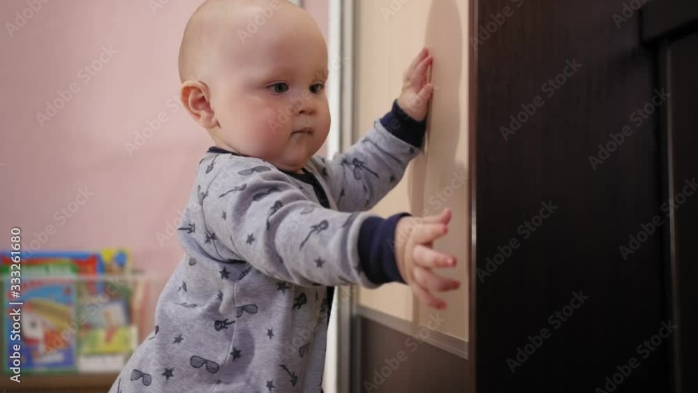 Cute nine-month-old baby standing near the cupboard and tapping it. Side view. Cute nine-month-old baby standing near the cupboard and tapping it. Side view.