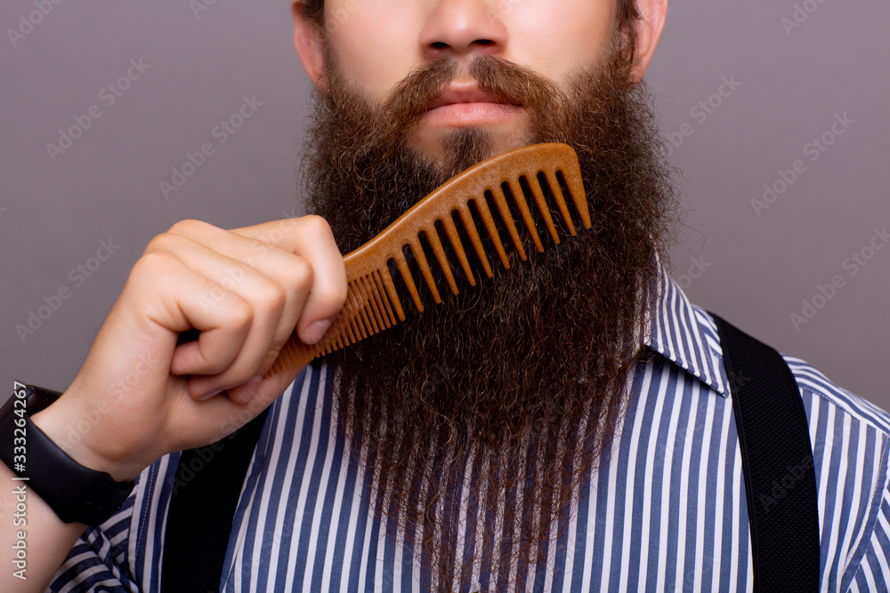 Closeup of a young man styling his long beard with a comb while standing alone in a studio against a gray background.